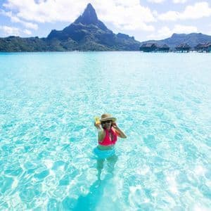 Eine Frau mit Hut und pinkfarbenem Bikini steht in klarem türkisfarbenem Wasser, hält ein Getränk, mit einem Berg und Überwasser-Bungalows hinter sich.