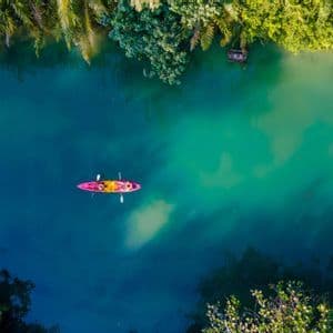 Luftaufnahme von zwei Personen, die ein rosa Kajak auf einem türkisfarbenen und blauen Fluss paddeln, umgeben von üppig grünen Bäumen.
