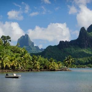 Ein kleines Boot schwimmt auf ruhigem Wasser unweit eines Palmenstrandes, dahinter erheben sich steile, grüne Berge vor einem blauen, bewölkten Himmel.