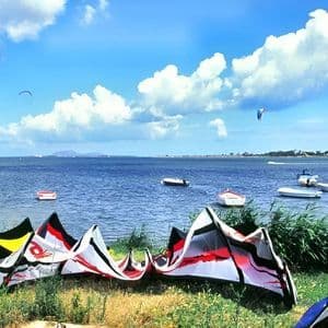 Cometas de kitesurf yacen en una orilla cubierta de hierba junto a una bahía con barcos y kitesurfistas a lo lejos bajo un cielo azul nublado.