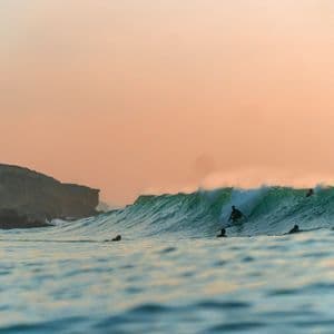 Viaggio di gruppo WeRoad di surfisti nell'oceano, con un surfista su una grande onda verde vicino a scogliere al tramonto.