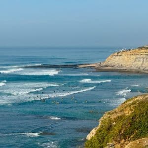 Surfisti sulle loro tavole sono sparsi su un oceano turchese con onde bianche che si infrangono vicino a un'alta scogliera sabbiosa.