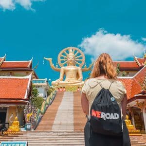 Una mujer con una mochila WeRoad mira hacia una larga escalera que conduce a una gran estatua de Buda dorado en un complejo de templos.