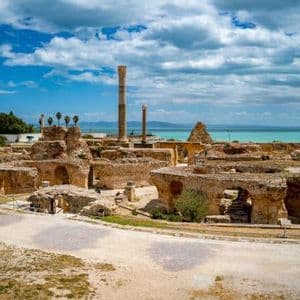 Antiche rovine in pietra e colonne di un sito archeologico si ergono in riva al mare turchese sotto un cielo azzurro parzialmente nuvoloso.