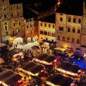 Una vista aerea di una piazza affollata di notte con bancarelle del mercato ed edifici illuminati da proiezioni di luce colorate.