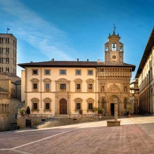 Una piazza in pietra vuota e illuminata dal sole, circondata da edifici storici, tra cui una chiesa, un campanile e una loggia ad archi, sotto un cielo azzurro.