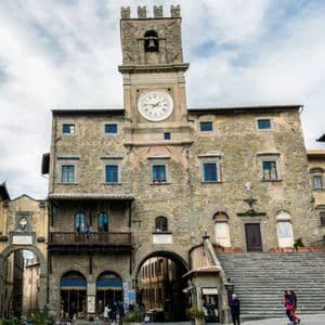 Un edificio storico in pietra con una grande torre dell'orologio e ampie scale si erge in una piazza europea sotto un cielo nuvoloso.