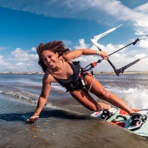 Una mujer sonriente en bañador negro hace kitesurf sobre el agua, inclinándose hacia atrás con una mano rozando la superficie bajo un cielo azul.