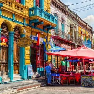 Una strada colorata con edifici gialli e blu brillanti accanto a un caffè all'aperto con tavoli e ombrelloni rossi.