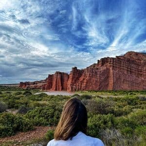 A WeRoad group leader seen from behind, looking out at a landscape of red rock formations and green shrubbery under a dramatic cloudy sky.