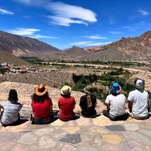 A WeRoad group trip sits with their backs to the camera on a stone viewpoint, looking out at a village in a vast mountain valley.
