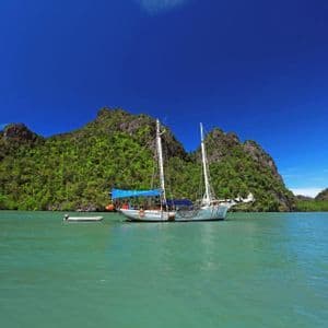 Ein zweimastiges Segelboot ankert in ruhigem, türkisfarbenem Wasser vor üppigen, grünen, felsigen Inseln.