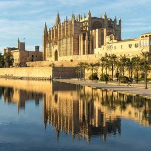 Una grande cattedrale gotica e mura in pietra che si riflettono in acqua calma, con palme lungo il lungomare.