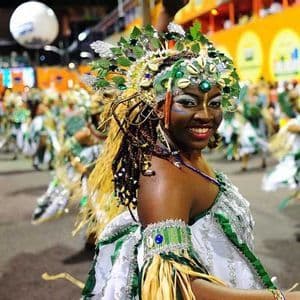 Une femme vêtue d'un costume de carnaval vert et blanc éclatant avec une grande coiffe sourit à l'appareil photo lors d'une parade nocturne.