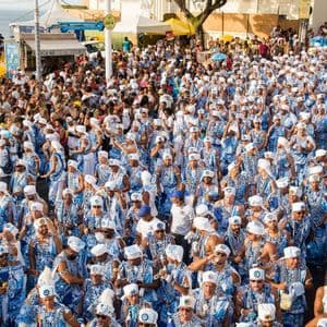 Un grand groupe de personnes en costumes assortis bleus et blancs défile dans une rue bondée près de la mer.