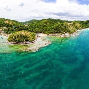 Une vue aérienne d'une île tropicale avec des collines verdoyantes, des eaux turquoise claires et une petite baie avec des bateaux amarrés.