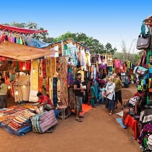Menschen schlendern an Ständen eines geschäftigen Außenmarktes vorbei, der unter einem strahlend blauen Himmel mit bunter Kleidung, Textilien und Taschen gefüllt ist.