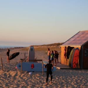 Un viaje en grupo WeRoad con tablas de surf en una playa de arena junto a una cabaña de surf de madera durante un atardecer dorado.