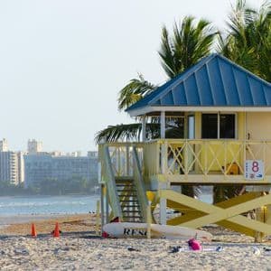 Una torre di salvataggio gialla con tetto blu si erge su una spiaggia sabbiosa, accanto a palme e con l'oceano sullo sfondo.