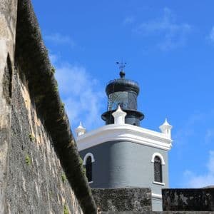 Una torre faro grigia con la cima nera vista da dietro un vecchio muro di pietra sotto un cielo blu.