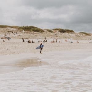 Un surfista con traje de neopreno sostiene una tabla de surf azul a la orilla del agua en una playa concurrida con dunas de arena bajo un cielo nublado.