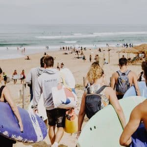 Un grupo de WeRoad con tablas de surf camina por un sendero arenoso hacia una playa concurrida con olas.