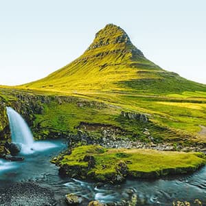 A wide-angle shot of a green, conical mountain with waterfalls cascading into a river in the foreground, under a clear sky.