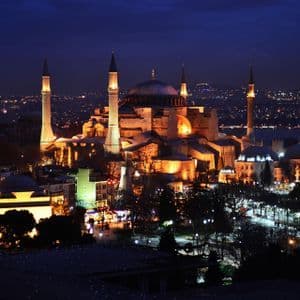 Una grande moschea con cupola e minareti illuminati da luci dorate di notte, sullo sfondo di un vasto skyline cittadino.