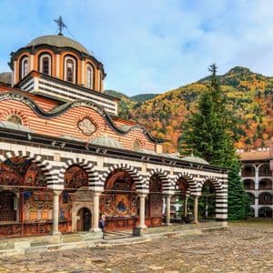 Un sontuoso monastero con archi a strisce e affreschi dettagliati in un cortile, e una montagna coperta di alberi autunnali sullo sfondo.