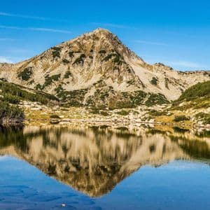 Una vetta rocciosa riflessa nelle acque calme e limpide di un lago alpino sotto un cielo azzurro e luminoso.