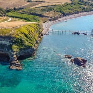 Veduta aerea di un pontile in legno che si protende da una costa rocciosa e rigogliosa verso un mare turchese, con dolci colline alle spalle.