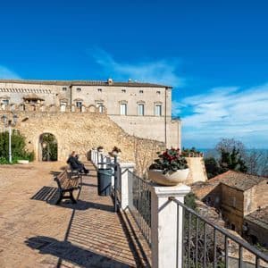 Un'ampia terrazza in mattoni con panchine si trova di fronte a un grande edificio in pietra, affacciata sul mare azzurro brillante sotto un cielo sereno.