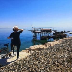 Una persona con un cappello bianco si trova con una bicicletta su una costa rocciosa, osservando un molo di legno per la pesca sul mare calmo e blu.