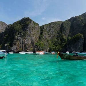 Hölzerne Longtail-Boote mit bunten Bändern liegen an einem Sandstrand im türkisfarbenen Wasser, mit einer großen Kalkstein-Karstinsel im Hintergrund.
