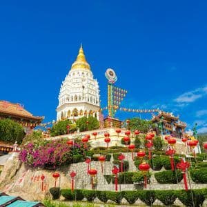 Ein großer Tempelkomplex mit einer weißen Pagode und einer goldenen Spitze, geschmückt mit roten Laternen, auf einem üppigen, terrassierten Hügel unter blauem Himmel.