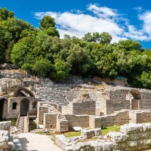 Ancient stone ruins of an amphitheater nestled into a hillside covered with green trees under a blue sky.
