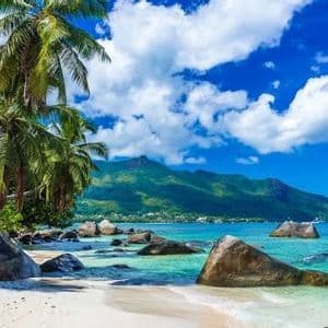 Palme rigogliose su una spiaggia di sabbia bianca con grandi massi, bagnata da acque cristalline turchesi, di fronte a una catena montuosa verde.