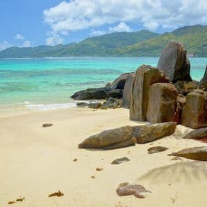 Grandi formazioni rocciose si ergono su una spiaggia sabbiosa accanto ad acque turchesi, con verdi colline visibili in lontananza sotto un cielo blu.