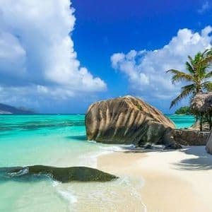 Grossi massi si trovano su una spiaggia di sabbia bianca accanto ad acque turchesi, con palme sotto un cielo parzialmente nuvoloso.