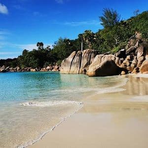 Onde leggere lambiscono una spiaggia di sabbia bianca accanto ad acqua turchese limpida, con grandi massi di granito e una lussureggiante collina verde sotto un cielo blu.
