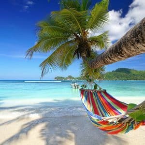 Un'amaca colorata pende da una palma su una spiaggia sabbiosa, con acqua turchese e un'isola verde sullo sfondo.