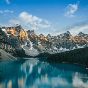 Sunlight hits the peaks of snow-capped mountains which reflect in a calm, turquoise lake surrounded by a pine forest.