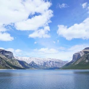 A wide, calm lake is flanked by steep, forested mountains under a blue sky with fluffy white clouds.