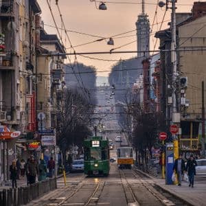 Veduta lungo una lunga via cittadina con tram sui binari, pedoni sui marciapiedi e una torre delle telecomunicazioni in lontananza.