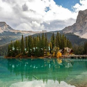 A wooden lodge on a forested peninsula is reflected in a turquoise alpine lake with mountains in the background.