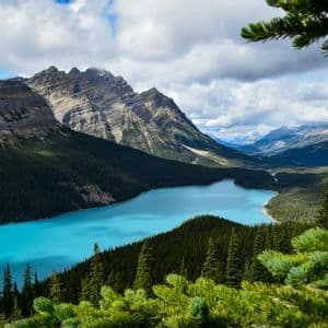 An elevated view of a bright turquoise lake surrounded by dense pine forests and rocky mountains under a cloudy sky.