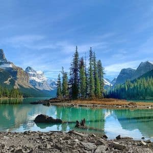 A small island with pine trees sits in a calm turquoise lake, surrounded by forests and snow-capped mountains.