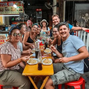 Eine WeRoad-Gruppe, die lächelnd an einem langen Tisch neben einem Streetfood-Stand Pad Thai mit Essstäbchen isst.