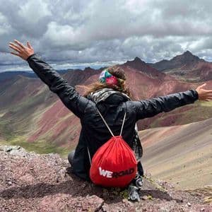 Un viajero con una mochila WeRoad roja se sienta en una cima rocosa, con los brazos extendidos, contemplando una colorida cordillera bajo un cielo nublado.