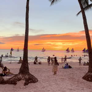 Menschen entspannen sich an einem Sandstrand mit Segelbooten auf dem Meer während eines lebhaften Sonnenuntergangs, umrahmt von Palmen.